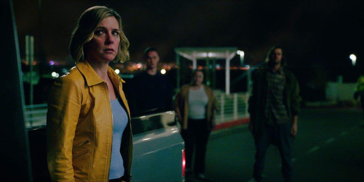 Carol in a yellow jacket standing apart from a group of hive members who are watching her from a distance in a night parking lot